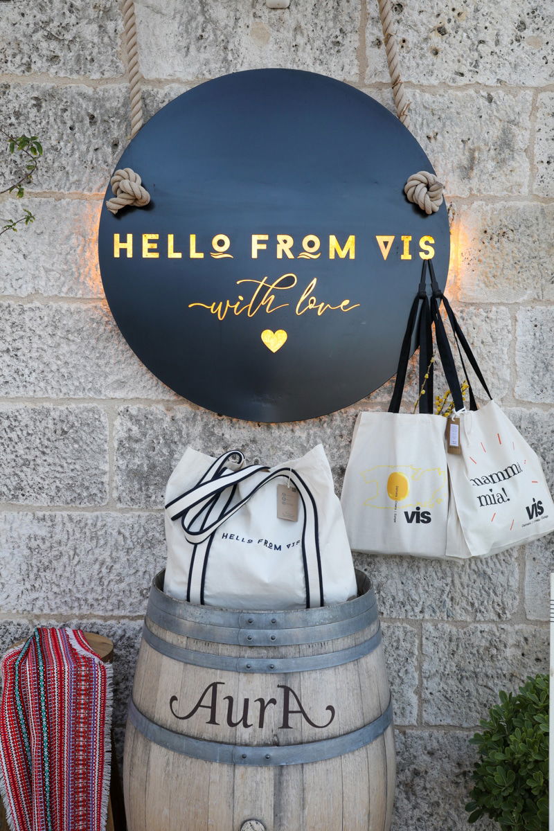 A sign reading "Hello from Vis with love" is hanging on a stone wall above a barrel labeled "Aura," with several tote bags, perfect for a Dubrovnik luxury cruise, hanging and displayed around it.