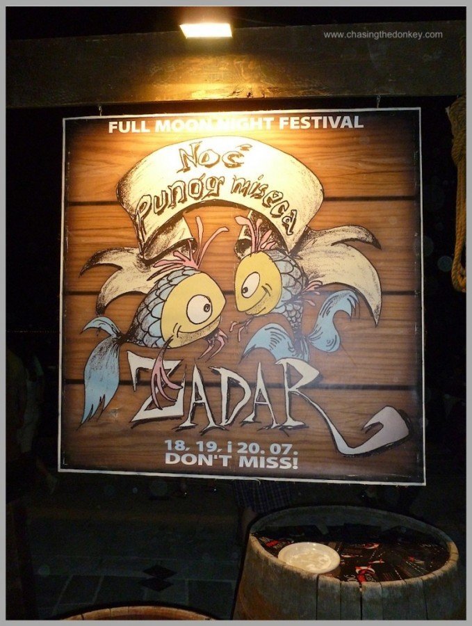 A festival sign reads "Full Moon Night Festival," featuring two cartoon fish and text "Nos Puno Miseca, Zadar, 18, 19, 20.07, Don't Miss!" with barrels underneath.