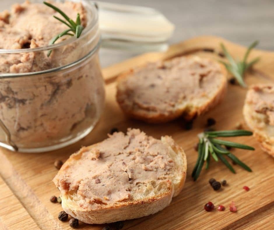 A jar of Pašteta Od Čvaraka (Pork Crackling Pâté) with a sprig of rosemary, slices of bread spread with pâté, and peppercorns on a wooden board.